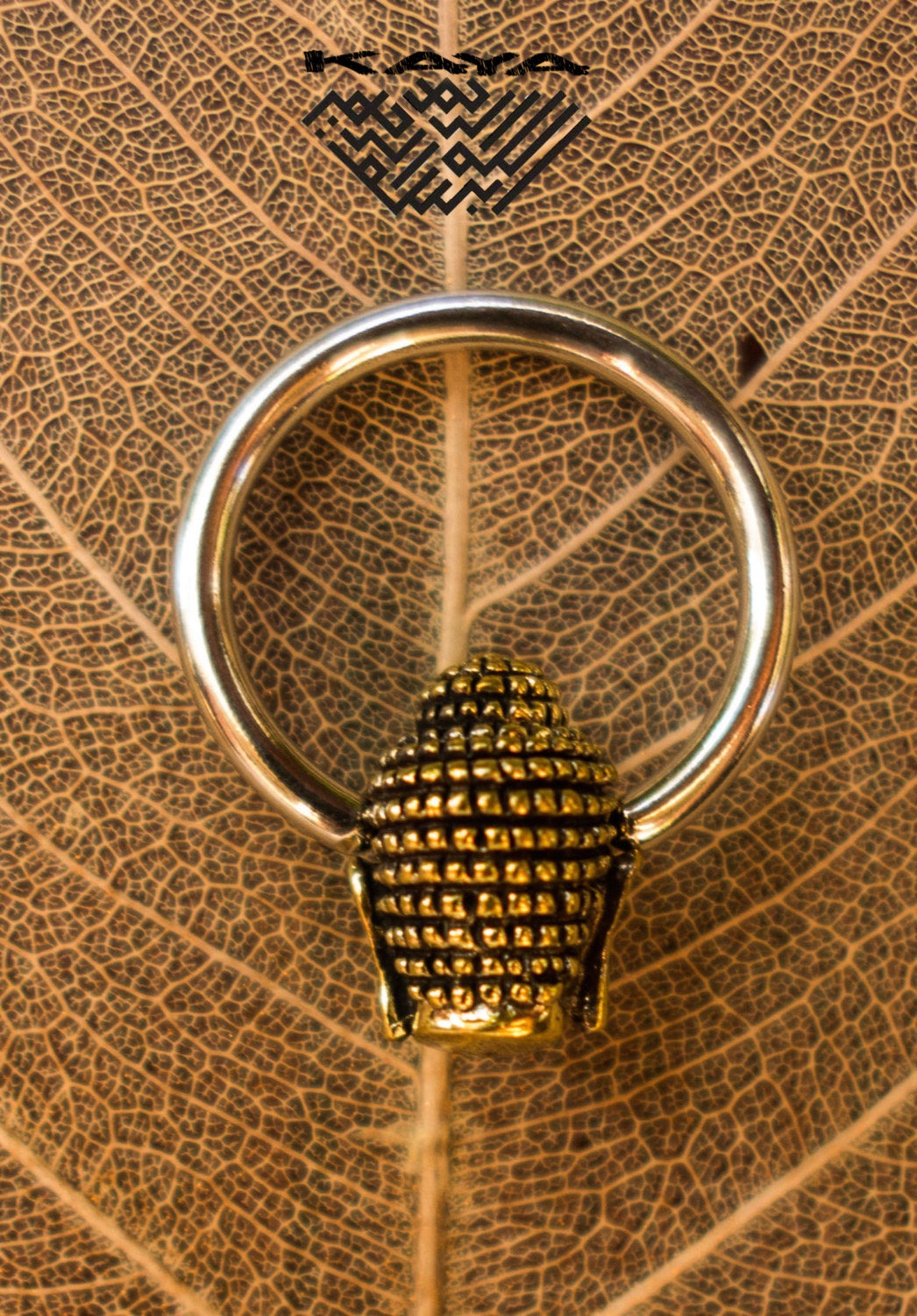 A gold and silver BUDDHA Silver Ring with Gold Pendant from Kaya Movement rests against an intricate, dried leaf with visible veins. The ring features a detailed small gold figurine resembling a beehive as its central element, reminiscent of an exquisite Buddha pendant design. Above the ring, a geometric design is faintly visible. Available in 18, 16 or 14 gauge sizes.